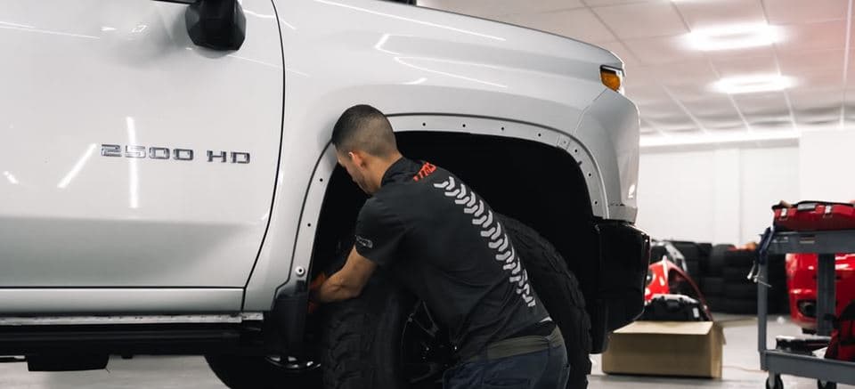 A technician fitting a wheel to a white Chevrolet Silverado 2500 HD inside the MAD Custom workshop