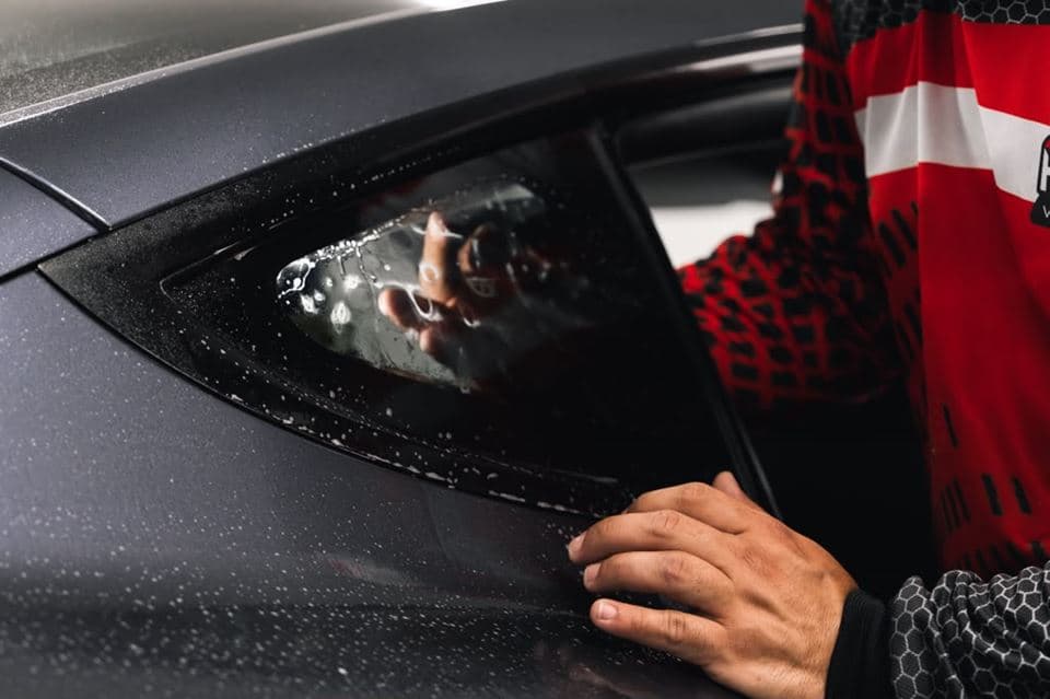 A MAD Custom installer squeegeeing window film onto the quarter glass of a dark grey car